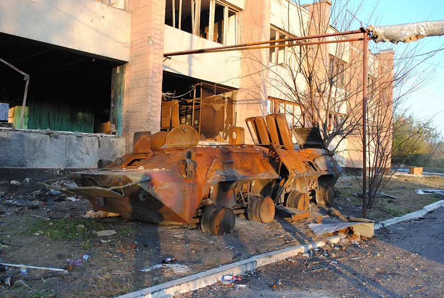 Ausgebranntes Kettenfahrzeug vor einem zerstörten Wohnhaus im Donbass.