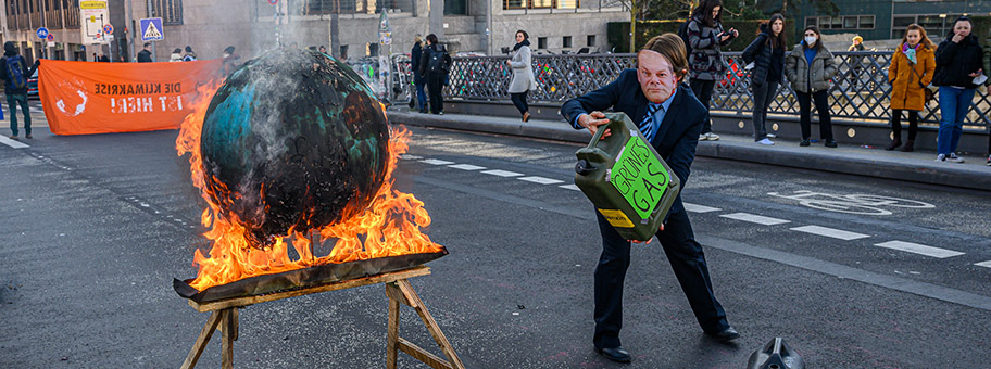 Extinction Rebellion zündet Weltkugel an, um auf IPCC-Bericht hinzuweisen. Aktivistin mit Scholz-Maske und Benzin-Kanister, auf dem Grünes Gas steht, giesst weiter Benzin ins Feuer. Marschallbrücke, Berlin, 28.02.2022.