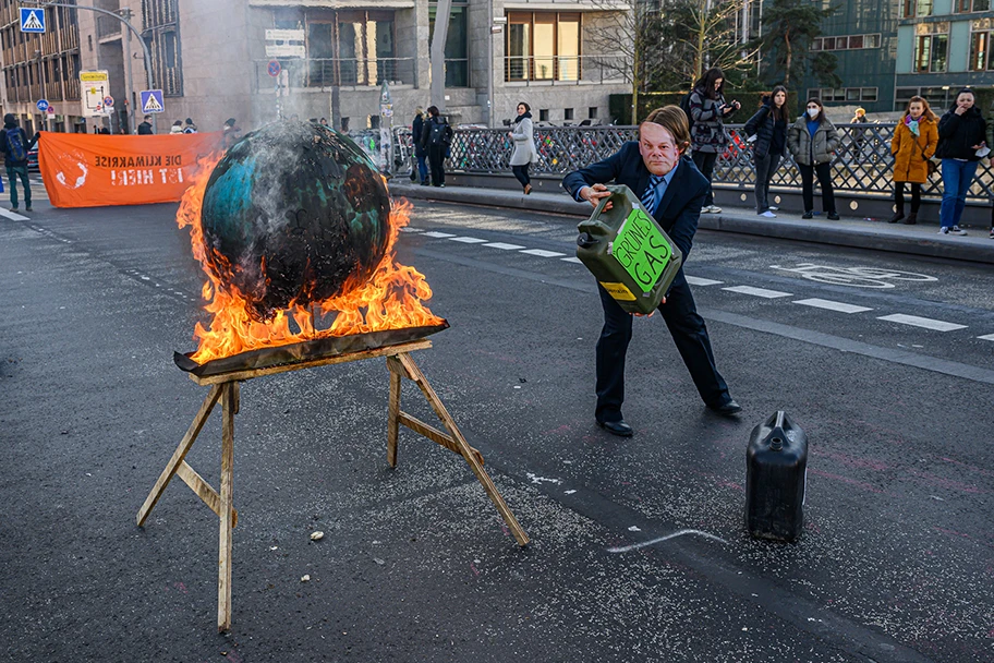 Extinction Rebellion zündet Weltkugel an, um auf IPCC-Bericht hinzuweisen. Aktivistin mit Scholz-Maske und Benzin-Kanister, auf dem Grünes Gas steht, giesst weiter Benzin ins Feuer. Marschallbrücke, Berlin, 28.02.2022.