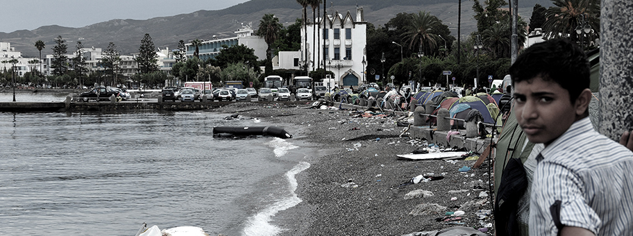 Ein Flüchtlingsjunge steht am Stadtstrand von Kos Stadt und schaut auf die Reste von gestrandeten Flüchtlingsbooten und die Zelte von Flüchtlingen.