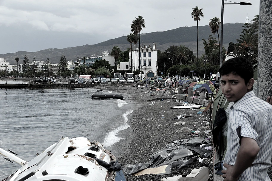 Ein Flüchtlingsjunge steht am Stadtstrand von Kos Stadt und schaut auf die Reste von gestrandeten Flüchtlingsbooten und die Zelte von Flüchtlingen.