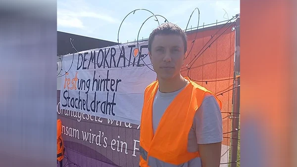 Simon Schwan vor dem Banner samt Stacheldraht am Zaun des Demokratiefests.